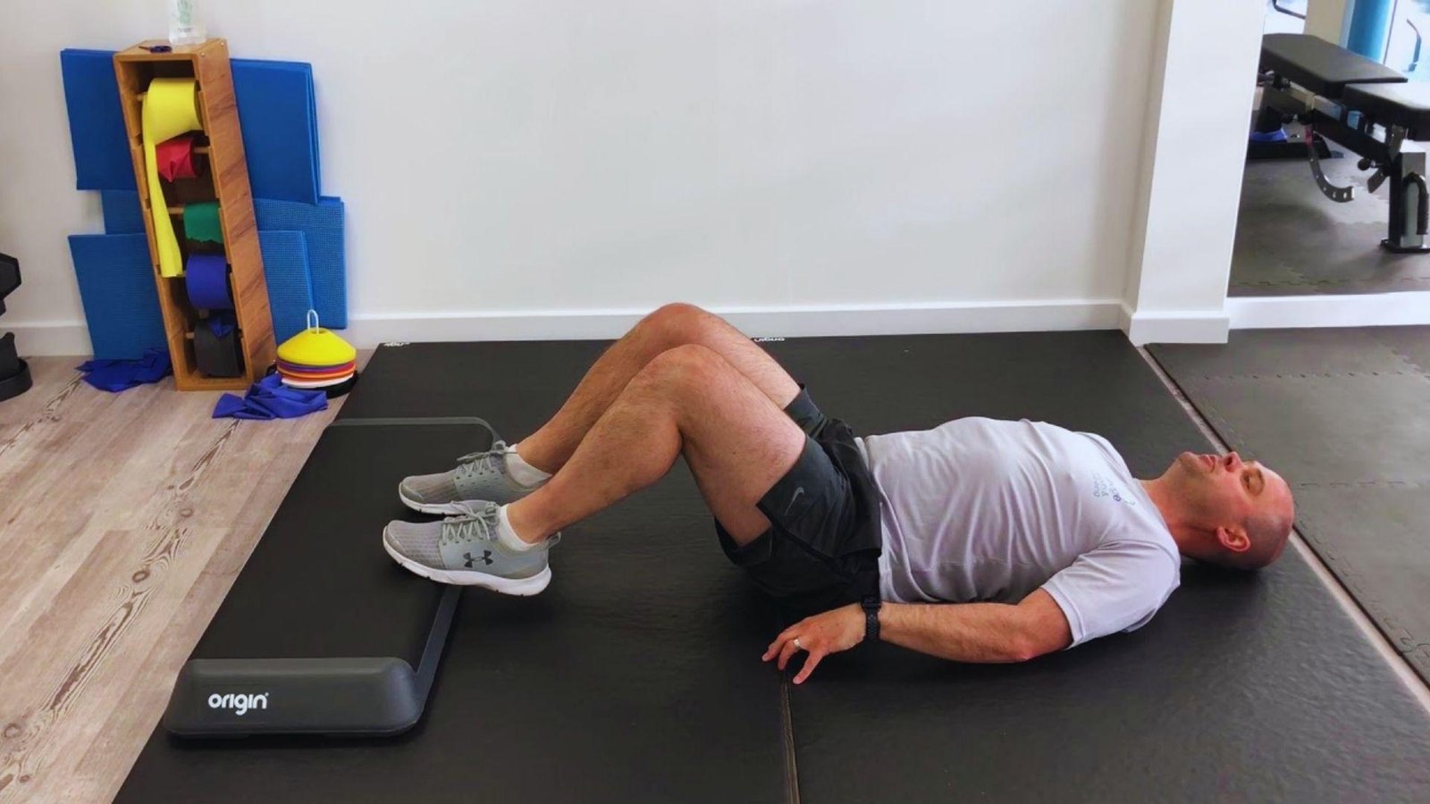 Man in gray shirt lies on mat with knees bent, heels on step platform, preparing for glute bridge