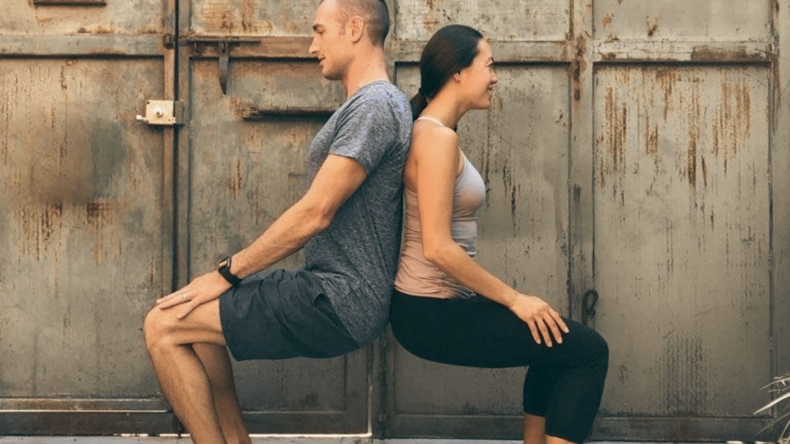 A man and woman sit facing each other on mats in a sunlit studio, legs lifted and soles touching.