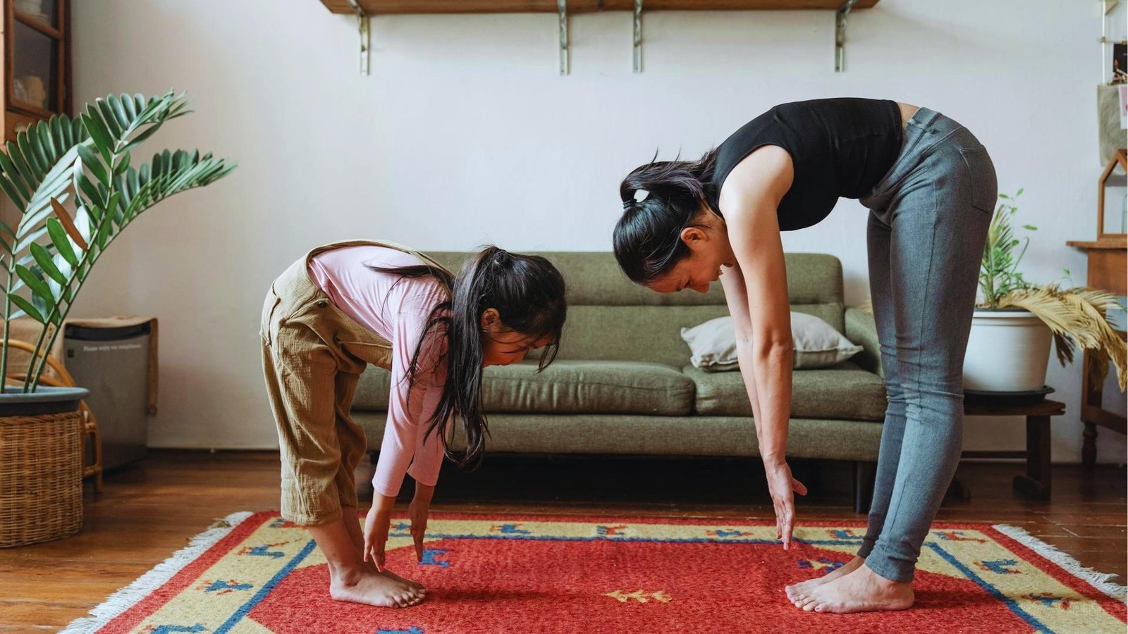 Mother and daughter bend forward in matching yoga pose on red rug, playfully reconnecting through movement at home.