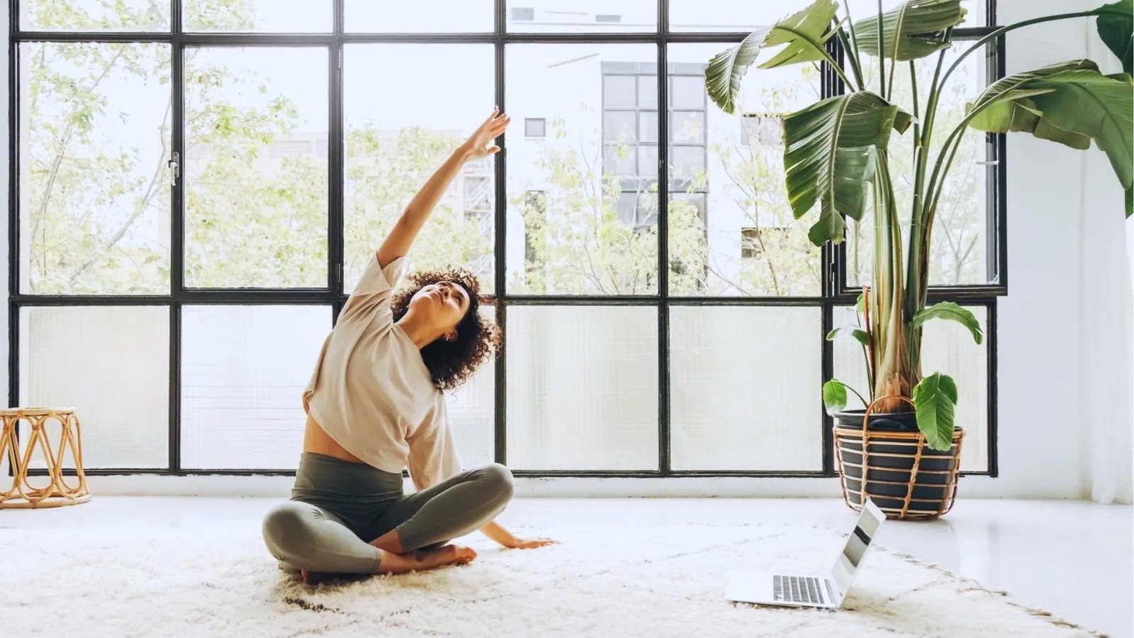 Woman in beige top and sage leggings does seated side bend on white rug, laptop nearby in sunlit room.