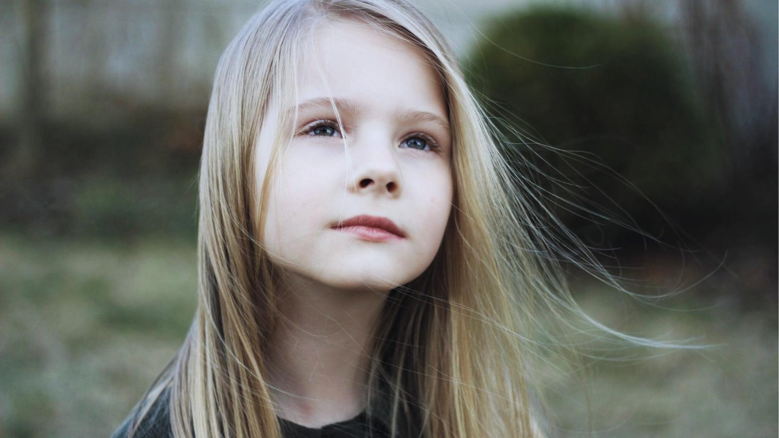 Young blonde girl gazes upward with wind in hair outdoors, expressing innocence, wonder, and inner child presence.