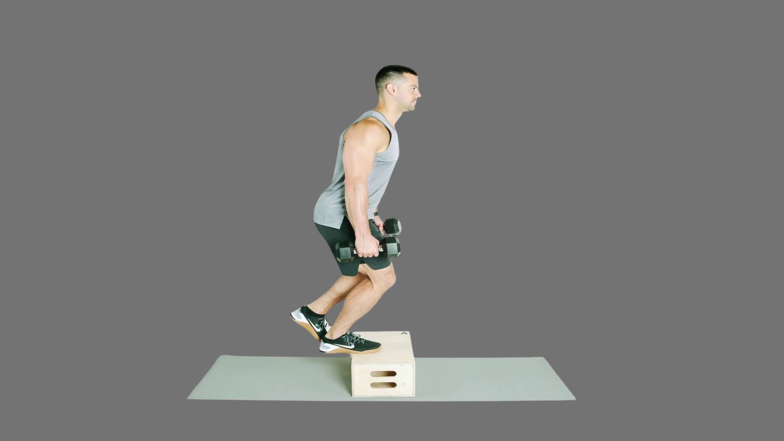 Man in gray tank top and black shorts performing weighted dumbbell box step-up on wooden plyo box