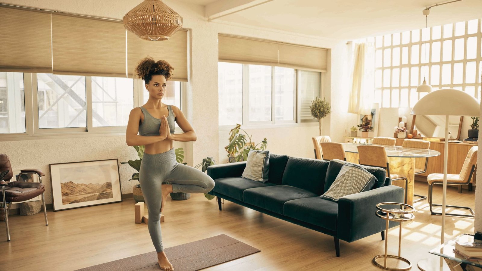 A woman balances in tree pose in a bright living room, palms together at her heart with steady focus.