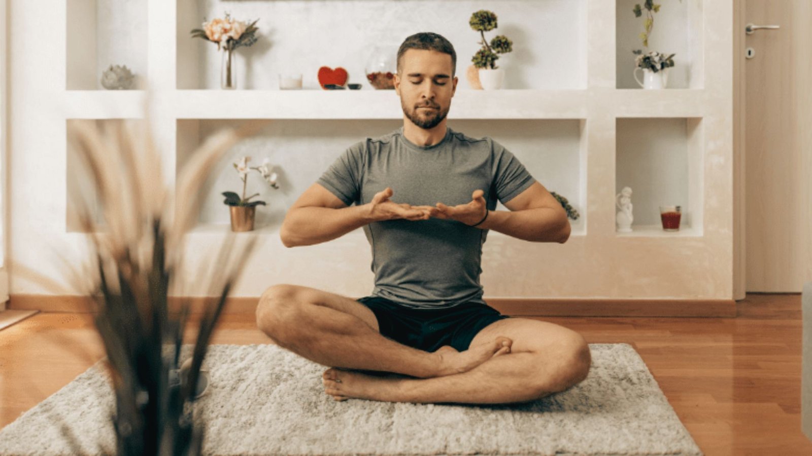 A man sits cross-legged on a rug in a meditative posture, hands hovering at his chest in mindful breathing.