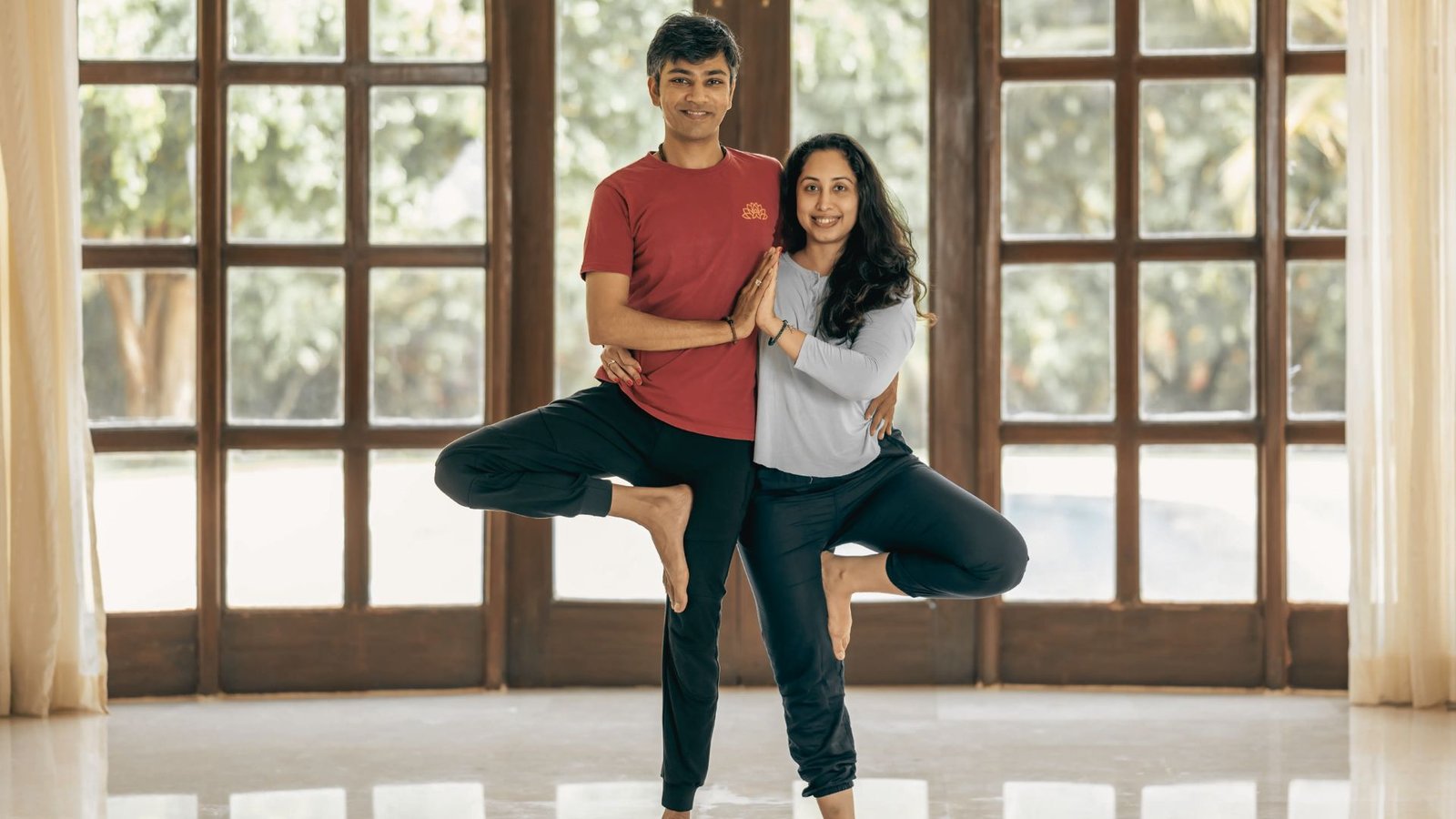 A couple sits back-to-back on a yoga mat in a calm studio with soft lighting.
