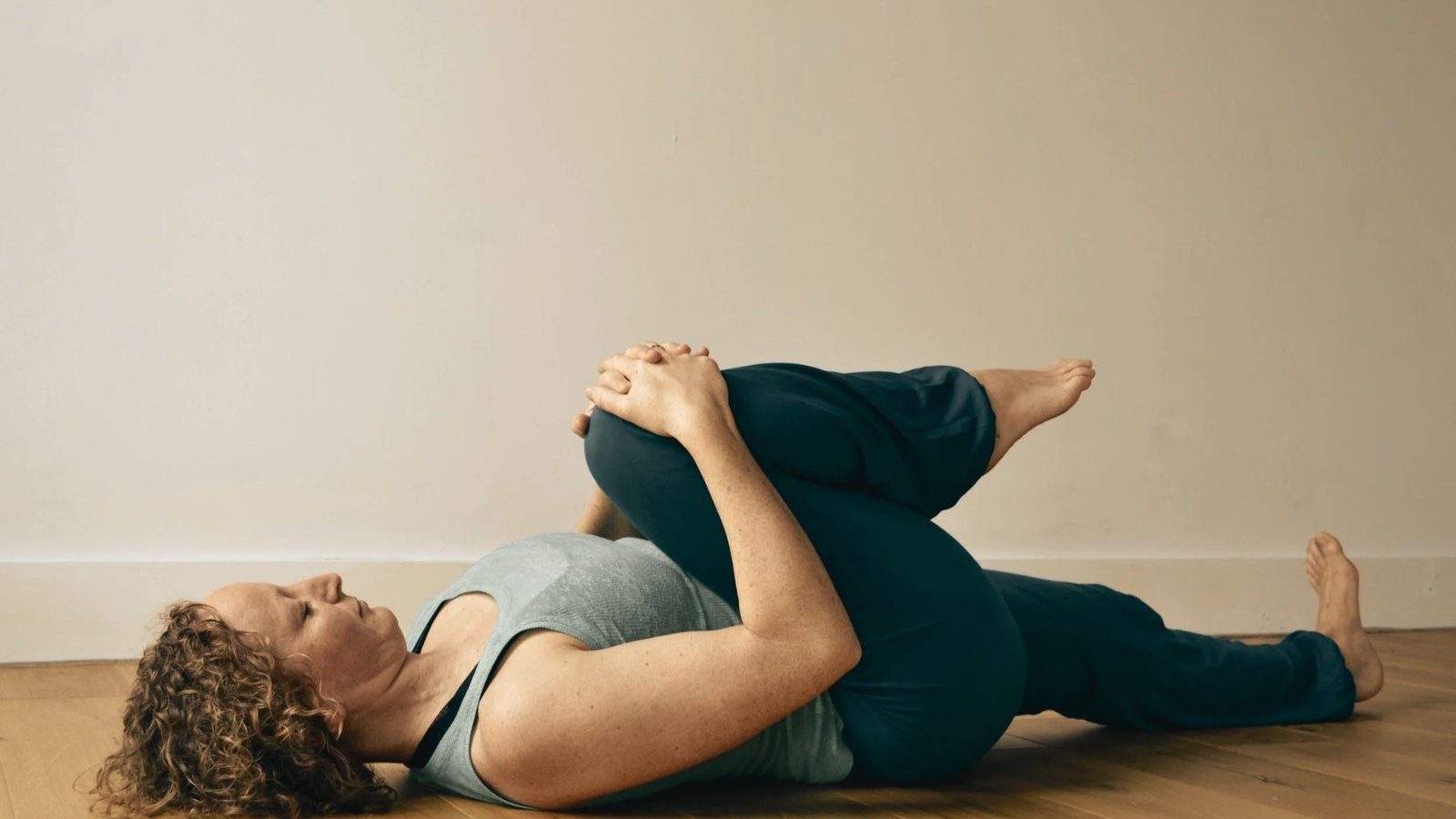 A woman lies on a wooden floor, hugging one knee to her chest in a gentle supine twist for somatic release.
