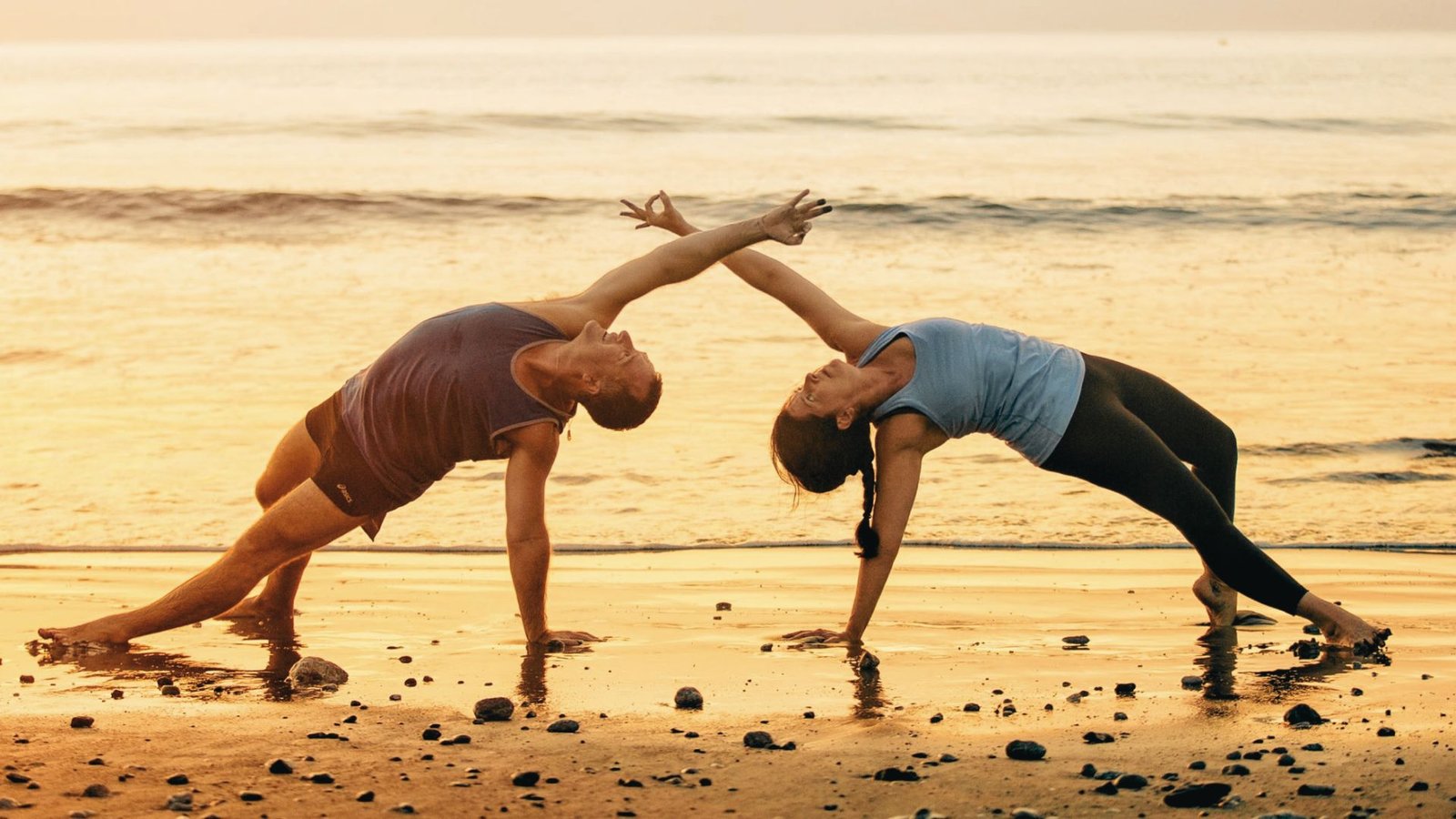 A man and woman mirror each other in Wild Thing Pose on a sandy beach at sunset, arms extended toward each other.