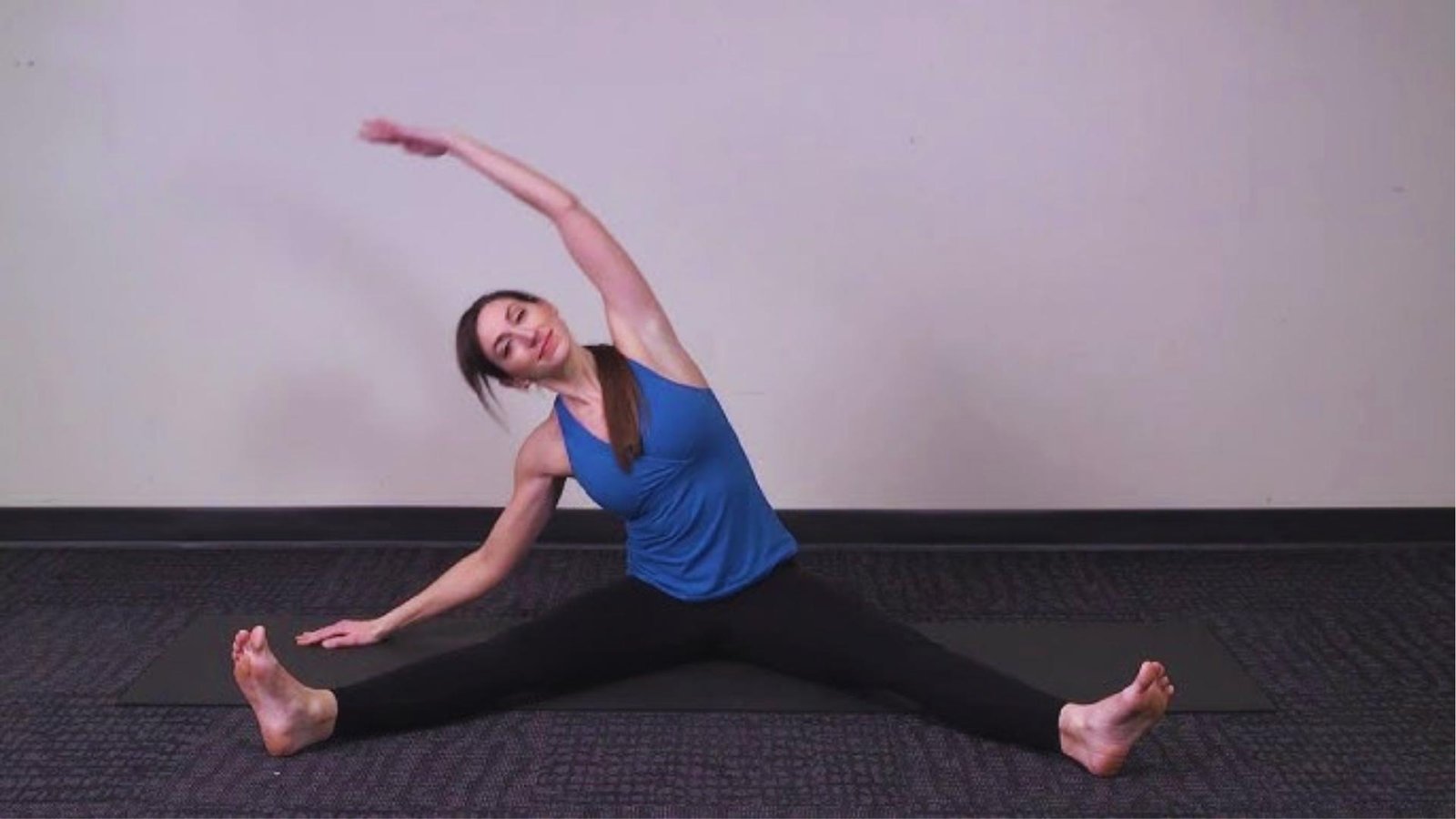 Woman in blue tank top does seated straddle side bend on mat, reaching arm overhead to stretch obliques and spine.