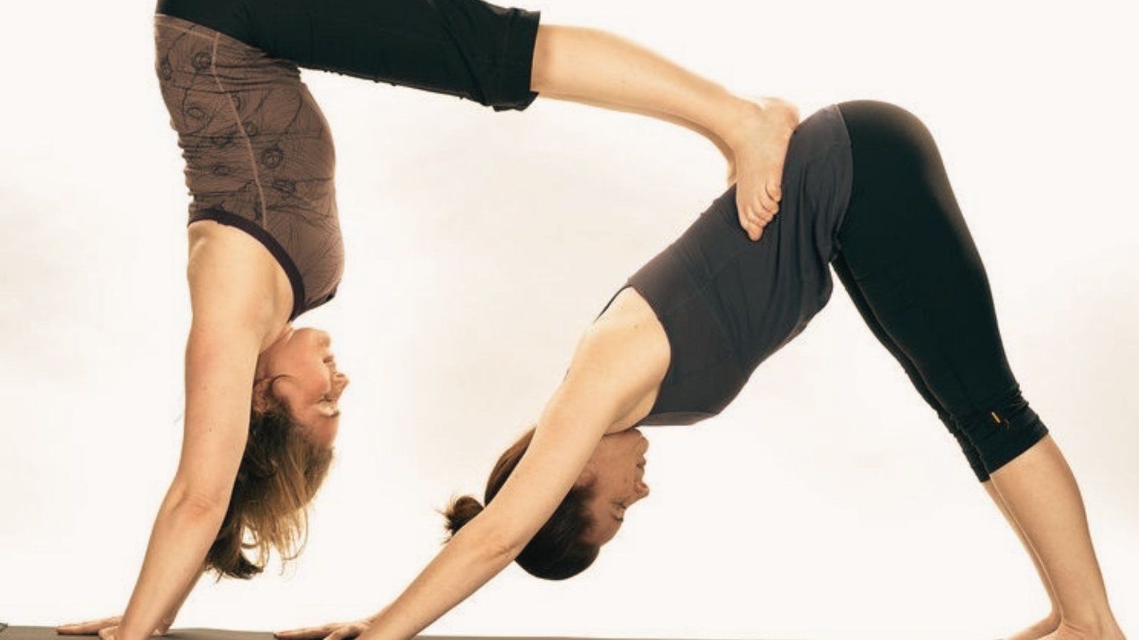 Two women practice partner yoga with one in Downward Dog and the other in a supported handstand on her back.